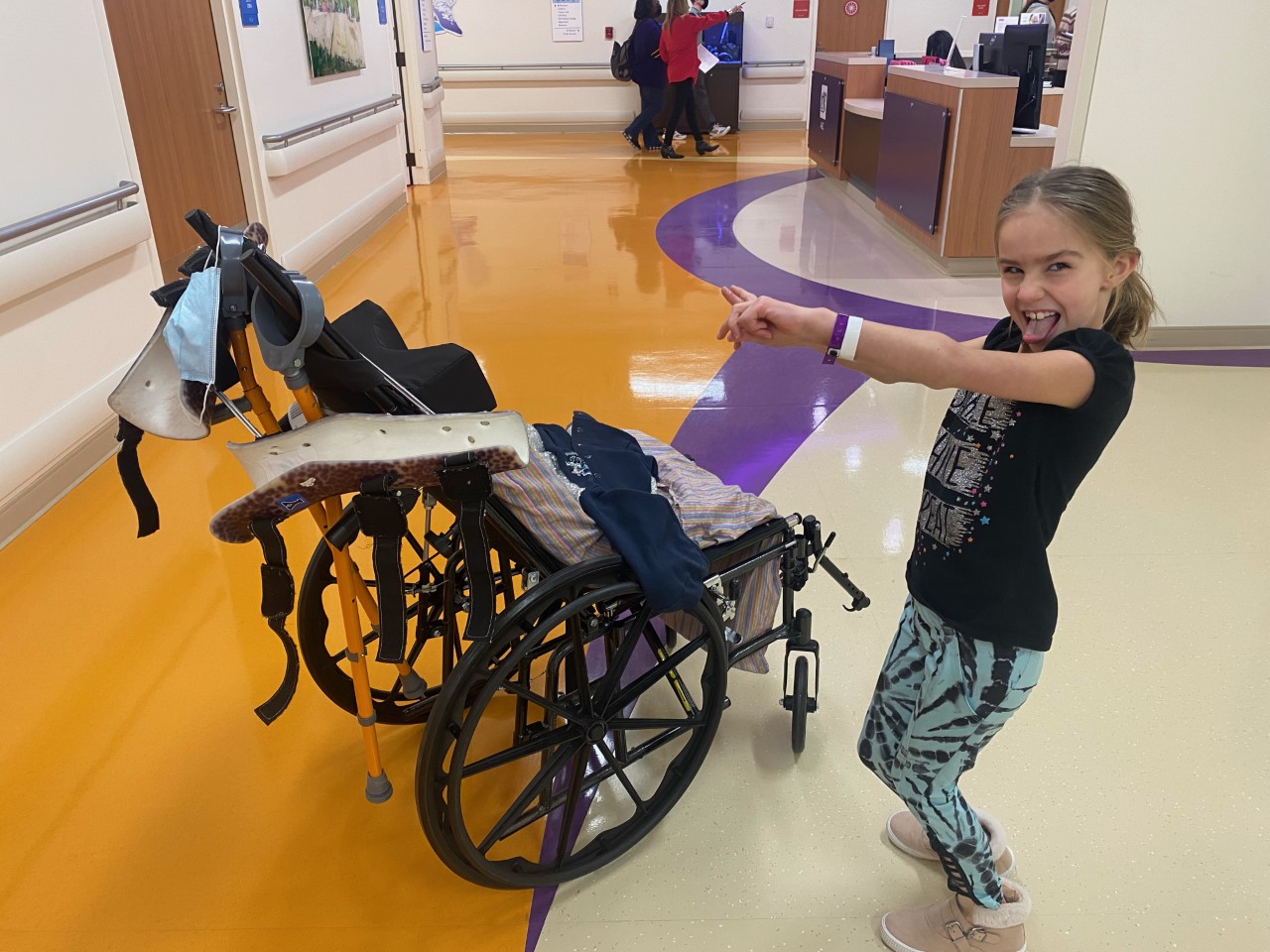 Smiling girl playfully points at an empty wheelchair in a hospital hallway