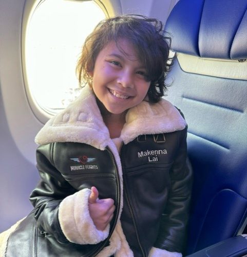 Smiling girl siting in an airplane, wearing a Miracle Flights' jacket