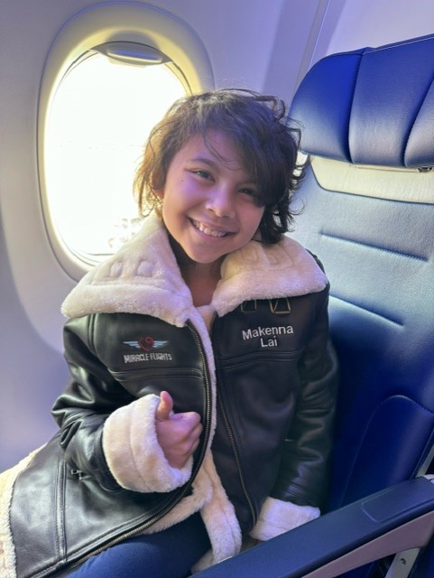Smiling girl siting in an airplane, wearing a Miracle Flights' jacket